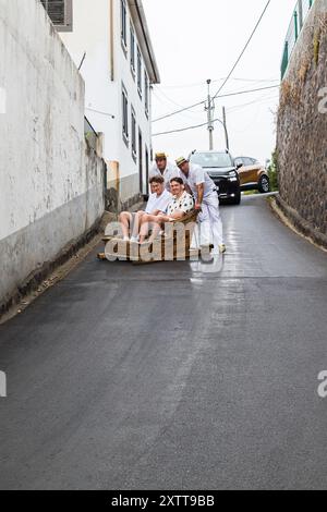 Zwei Touristen, die eine Fahrt mit Rodeln aus Camacha-Korb genießen, während sie von Monte nach Funchal, Madeira, fahren, gesehen am 31. Juli 2024. Stockfoto