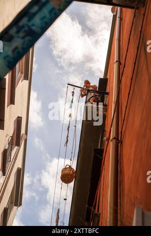 Neapel, Italien - 24. Juni 2024: Ein Mann steht auf einem Balkon und hebt einen Korb mit Seilen in einer engen städtischen Gasse mit hellem Tageslicht und bewölktem Himmel. Stockfoto