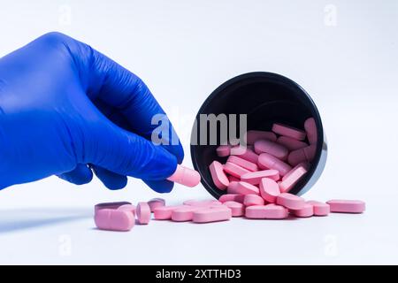 Doktor Hand in blauen Handschuhen, die verschüttete rosa Pillen aus einem schwarzen Plastikbehälter auf weißem Hintergrund aufnimmt. Stockfoto