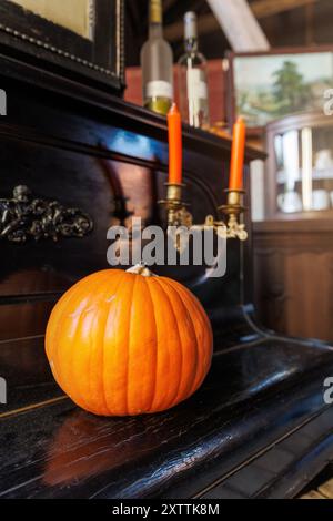 Großer reifer orangener Kürbis sitzt auf einem Vintage-schwarzen Klavier mit verzierten Details, begleitet von zwei angezündeten orangefarbenen Kerzen in antikem Halter. Die Einstellung Stockfoto