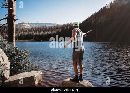 Mann fischt auf Felsen in See mit Bäumen und Bergen Stockfoto