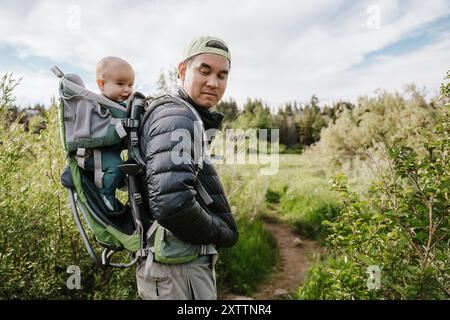 Asain Vater mit Rucksack, der ein lächelndes Baby in üppiger Landschaft ansieht Stockfoto