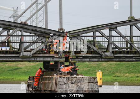 Elsfleth, Deutschland. August 2024. Die Arbeiter stellen sicher, dass die Brücke perfekt sitzt, damit die neue provisorische Brücke beim Anheben an ihrem Platz ist. Der „Athlet“-Schwimmkran bringt den 24 Meter langen und 58 Tonnen schweren Brückenabschnitt als Ersatz für die Eisenbahnbrücke, die einige Wochen zuvor von einem Tankfahrzeug gerammt wurde. Diese Brücke war auch eine provisorische Brücke, da bereits im Februar ein Lastkahn auf die reguläre Eisenbahnbrücke traf. Die Brücke und die Freileitungen wurden so stark beschädigt, dass der Zugverkehr eingestellt werden musste. Quelle: Lars Penning/dpa/Alamy Live News Stockfoto