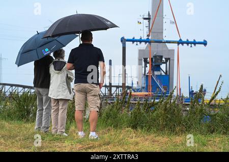 Elsfleth, Deutschland. August 2024. Die Familie Wallinger wirft trotz Regen einen Blick auf die Brückenbaustelle. Der „Athlet“-Schwimmkran bringt den 24 Meter langen, 58 Tonnen schweren Brückenabschnitt als Ersatz für die Eisenbahnbrücke, die einige Wochen zuvor von einem Tankfahrzeug gerammt wurde. Diese Brücke war auch eine provisorische Brücke, da bereits im Februar ein Lastkahn auf die reguläre Eisenbahnbrücke traf. Die Brücke und die Freileitungen wurden so stark beschädigt, dass der Zugverkehr eingestellt werden musste. Quelle: Lars Penning/dpa/Alamy Live News Stockfoto