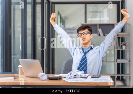 Müder und überarbeiteter junger Geschäftsmann, der seine Arme vor Erschöpfung ausstreckt, während er spät im Büro arbeitet Stockfoto