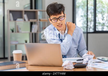 Müder und überarbeiteter junger Geschäftsmann, der seine Hand benutzt, um seinen Nacken zu massieren, während er spät im Büro arbeitet Stockfoto