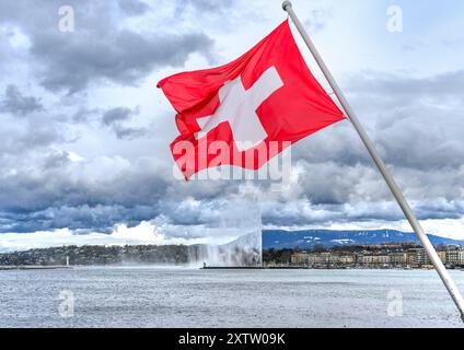 Die markante rote Schweizer Flagge hängt über dem kristallklaren Wasser des Genfer Sees und dem berühmten Brunnen (Jet d'Eau de Genève) in der Ferne. Stockfoto