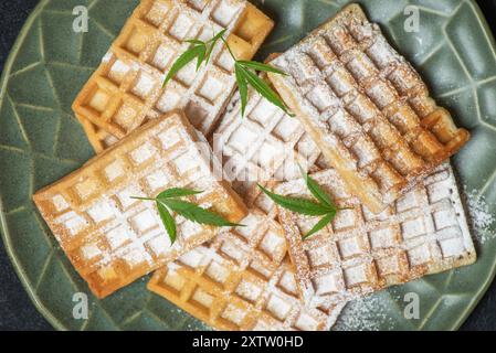 Köstliche Waffeln mit Puderzucker bestreut und mit frischen Marihuanablättern auf einem grünen Teller dekoriert. Nahaufnahme, Draufsicht Stockfoto