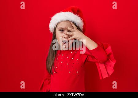 Entzückendes hispanisches Mädchen im Schock! Über die Finger schauen, Gesicht und Augen mit der Hand bedecken, Angst im Auge, während sie weihnachtsmütze tragen. wow, die isolierte Re Stockfoto