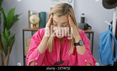 Eine gestresste junge blonde Frau, die ein rosa Hemd trägt, hat Kopfschmerzen in ihrem überfüllten Büro Stockfoto