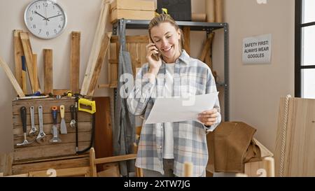 Eine lächelnde junge Frau telefoniert am Telefon, während sie in einer geschäftigen Holzwerkstatt Pläne hält. Stockfoto