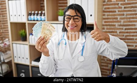 Die hispanische Ärztin hält brasilianisches Geld mit Daumen nach oben in einem Klinikinnenraum. Stockfoto