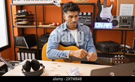 Hübscher hispanischer Mann, der eine Akustikgitarre in einem modernen Musikstudio mit orangefarbenen Details spielt. Stockfoto