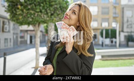 Eine zufriedene hispanische Frau hält kolumbianische Pesos im Freien in einem Stadtpark und stellt Finanzen und Lebensstil zur Schau. Stockfoto