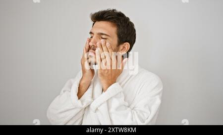 Ein junger hispanischer Mann in einem weißen Gewand berührt sein Gesicht und vermittelt eine Hautpflege- oder Pflegeroutine vor einem isolierten weißen Hintergrund. Stockfoto