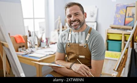 Hübscher hispanischer reifer Mann mit grauen Haaren in einem Atelier, mit Schürze und Pinsel. Stockfoto