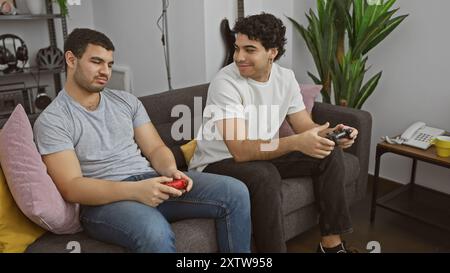 Zwei Männer spielen gemeinsam Videospiele und genießen die Freizeit drinnen, während sie bequem auf einem grauen Sofa sitzen. Stockfoto