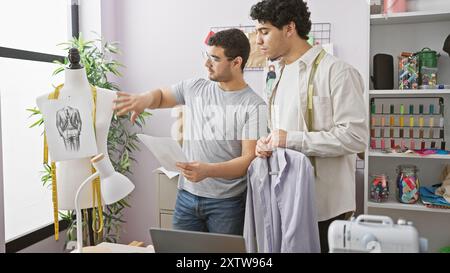 Zwei Männer analysieren ein Kleidungsdesign in einer Schneiderei, umgeben von Nähzubehör und bunten Garnen. Stockfoto