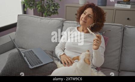 Eine reife Frau mit lockigen Haaren sitzt drinnen in einem Wohnzimmer mit einer Brille, begleitet von ihrem chihuahua-Hund und arbeitet an einem Laptop. Stockfoto
