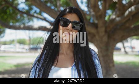 Attraktive brünette Frau in Sonnenbrille und lässigem Outfit, die in einem sonnigen Park in der Stadt steht. Stockfoto