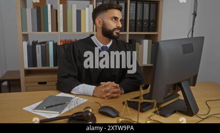 Hübscher junger hispanischer Mann mit einem Bart im Richtermantel, der im Büro sitzt, mit Computer und Rechtsdokumenten, die durchdacht aussehen Stockfoto