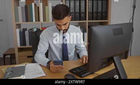 Hübscher junger hispanischer Mann mit Bart, der ein Smartphone am Schreibtisch in einem modernen Büro-Innenarbeitsplatz benutzt Stockfoto