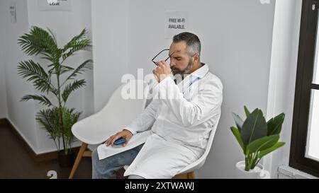 Ein reifer bärtiger Mann in einem weißen Mantel benutzt das Smartphone, während er in einem modernen Warteraum mit Pflanzen sitzt. Stockfoto