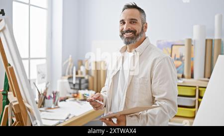 Ein bärtiger älterer hispanischer Mann mit einem sanften Lächeln hält eine Palette und einen Pinsel im Atelier eines Künstlers und verkörpert Kreativität und Erfahrung. Stockfoto
