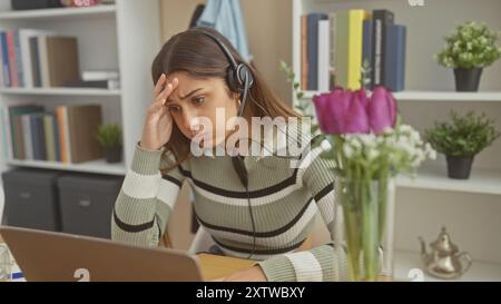 Eine gestresste hispanische Frau mit einem Headset schaut in einem modernen Home Office mit Büchern und Pflanzen auf einen Laptop. Stockfoto