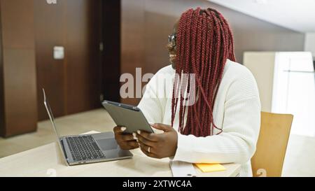 afroamerikanische Geschäftsfrau mit Zöpfen lächelt in einem Büro, hält ein Tablet und sitzt neben einem Laptop. Stockfoto
