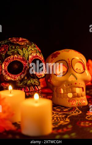 Vertikale Komposition mit zwei Schädeln auf einer Tischdecke zum Tag der Toten: Einer mit metallischen Blumen, der andere mit einer beleuchteten Kerze. Ma Stockfoto