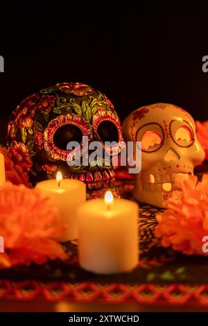 Vertikale Komposition mit zwei Schädeln auf einer Tischdecke zum Tag der Toten: Einer mit metallischen Blumen, der andere mit einer beleuchteten Kerze. Ma Stockfoto