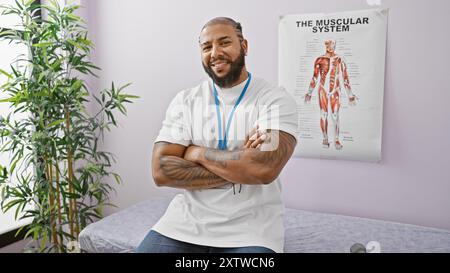 Lächelnder bärtiger Mann mit Tattoos und Schlüsselband, der in einer Klinik steht, mit einem muskulären System-Poster im Hintergrund. Stockfoto