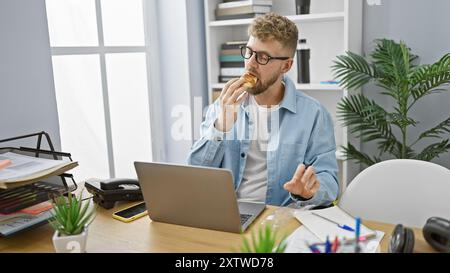 Hübscher junger Mann mit Bart, der in einem unordentlichen Büro isst, umgeben von Technologie und Pflanzen. Stockfoto