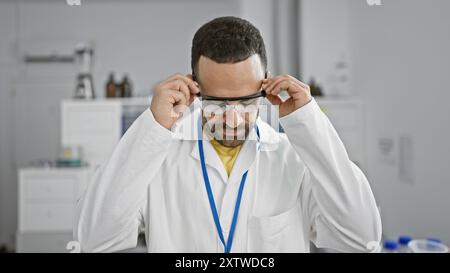 Ein junger bärtiger hispanischer Mann passt eine Schutzbrille in einem weißen Labormantel an. Stockfoto