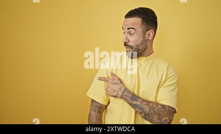 Hübscher hispanischer Mann mit Bart und Tätowierungen, die zur Seite zeigen, auf einem gelben isolierten Hintergrund Stockfoto