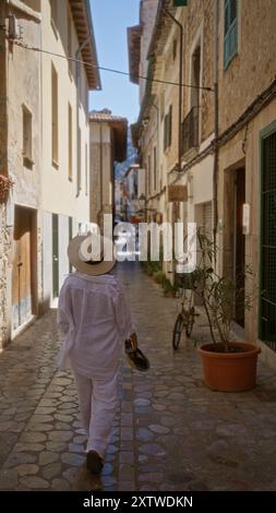 Frau, die in einer malerischen, gepflasterten Straße in fornalutx, mallorca, spanien, spaziert, in einem weißen Outfit und Hut an einem sonnigen Tag und zeigt das charmante Stockfoto