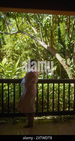 Eine ruhige Frau genießt eine Tasse Kaffee auf einem Balkon mit Blick auf das üppige Grün in einem bali Resort. Stockfoto