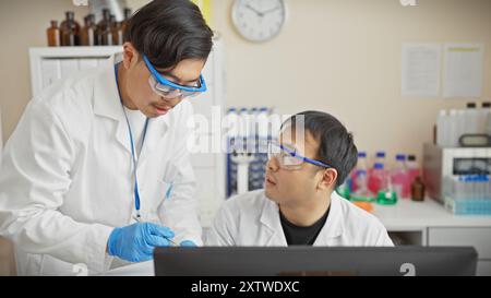 Zwei männliche Wissenschaftler arbeiten in einem Labor zusammen, tragen eine Schutzbrille und diskutieren über einen Computer. Stockfoto