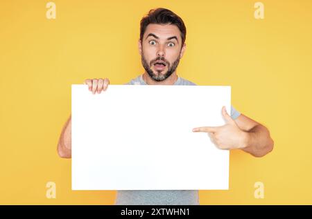 Ein Typ mit leerer Bannerwerbung, isoliert im Studiohintergrund. Ein Mann hält ein leeres Poster mit einem leeren Poster, das mit dem Finger zeigt, und einem Gesten-Index Stockfoto