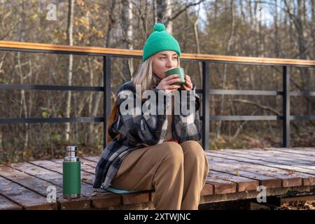 Eine ruhige, entspannte Frau, die sich mit heißem Tee aufwärmt, macht eine Pause auf dem ökologischen Weg im Herbstwald Stockfoto