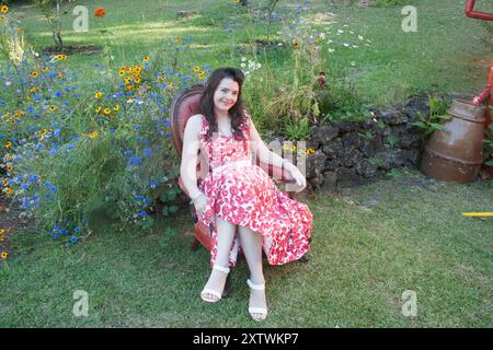 Lächelnde Frau, die auf einem roten Stuhl in einem Garten sitzt, mit Wildblumen in einem weiß-roten Blumenkleid und weißen Sandalen. Stockfoto