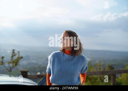 Die lächelnde Frau in einem blauen Pullover und einem orangefarbenen Hemd steht draußen mit einem malerischen Blick im Hintergrund. Stockfoto