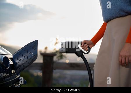 Nahaufnahme einer Person, die während des Sonnenuntergangs an einer Ladestation ein Ladegerät für Elektrofahrzeuge an einem Auto anschließt. Stockfoto