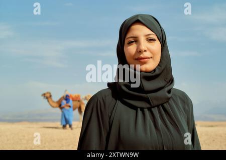 Eine lächelnde Frau in einem Hijab steht in einer Wüstenlandschaft mit einem Kamel und einer Person in blauer traditioneller Kleidung im Hintergrund. Stockfoto