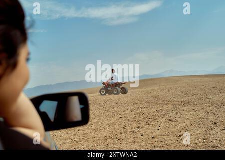 Eine Frau beobachtet einen Mann auf einem Quad in einer kargen Landschaft, von einem Autospiegel aus gesehen. Stockfoto