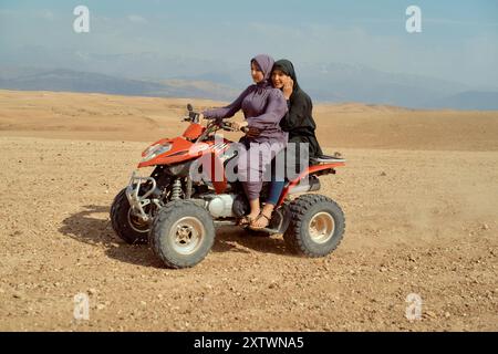 Zwei junge Mädchen lächeln, während sie auf einem roten Quad sitzen, in einer Wüstenlandschaft mit Bergen im Hintergrund. Stockfoto