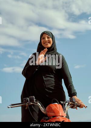 Lächelnde Frau in einem Hijab, die einen roten Roller vor einem blauen Himmel fährt. Stockfoto