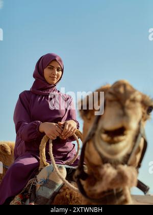 Eine lächelnde Frau in einem violetten Hijab sitzt auf einem Kamel, ein anderes Kamel lächelt im Vordergrund. Stockfoto