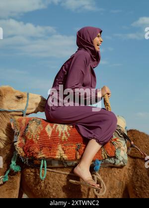 Lächelnde Frau in einem Hijab, der auf einem Kamel unter klarem blauen Himmel reitet. Stockfoto